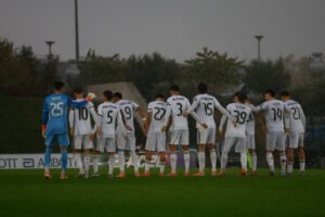Jugadores del Real Madrid Castilla alineados de espaldas guardando un minuto de silencio bajo la lluvia.